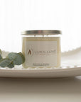 A three-wick clear glass candle with natural ivory wax sitting on a white hobnail glass tray. On the left is a sprig of eucalyptus leaves.