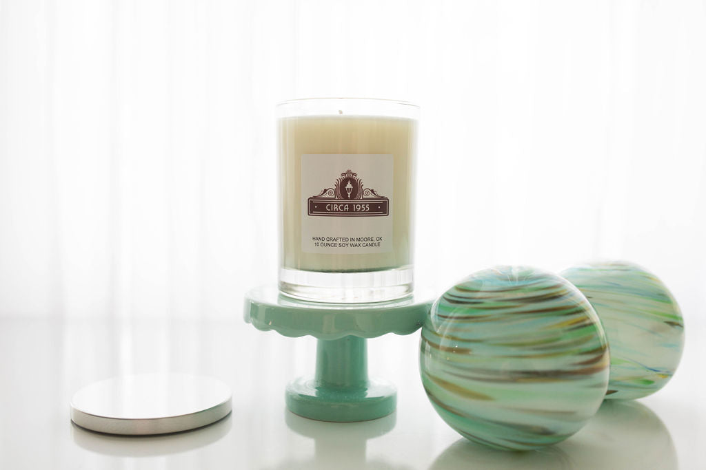 A clear glass candle with natural ivory wax sitting on a mini cake tray that is light blue.  On the right is a decorative glittery orb with blue, turquoise and gold swirls.