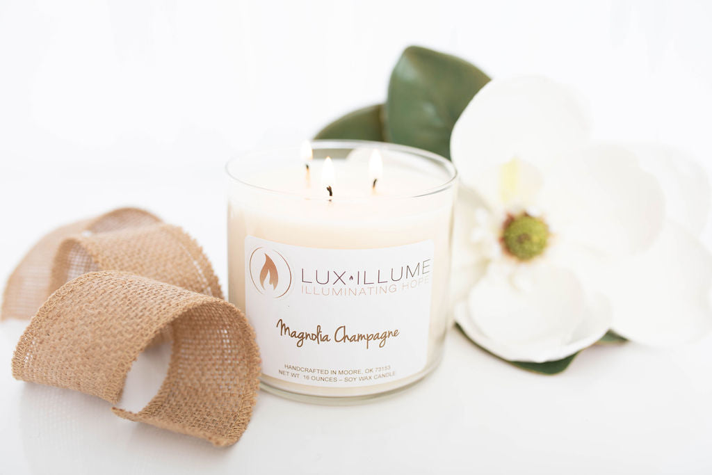 A three-wick clear glass candle with natural ivory wax.  On the left is a burlap ribbon and on the right is a magnolia blossom.
