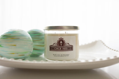 A three-wick clear glass candle with natural ivory wax sitting on a white hobnail glass tray.  On the left is two decorative glittery orbs with turquoise and gold swirls.