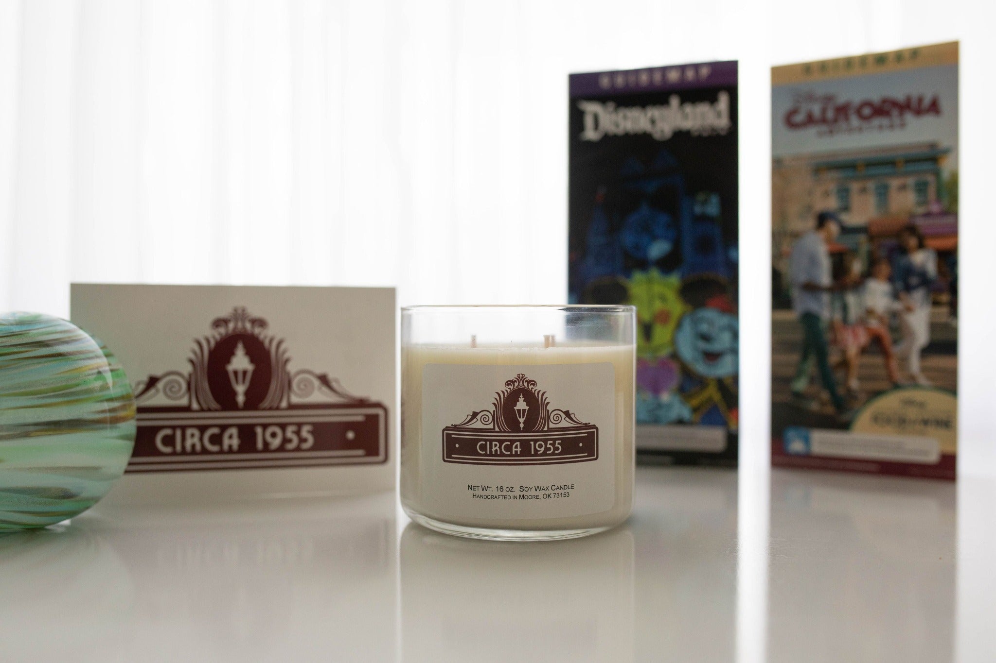 An overhead view of a single wick glass candle on a white hobnail tray with various Disney park maps arranged to the right.