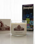 An overhead view of a single wick glass candle on a white hobnail tray with various Disney park maps arranged to the right.