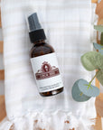 An amber glass bottle laying on top of a natural linen towel above a slatted wood bowl or tray.