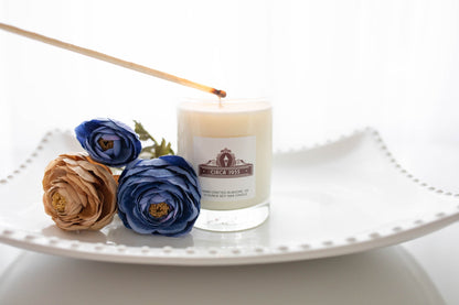 A single wick clear glass candle on a white hobnail tray with dried flowers next to it.