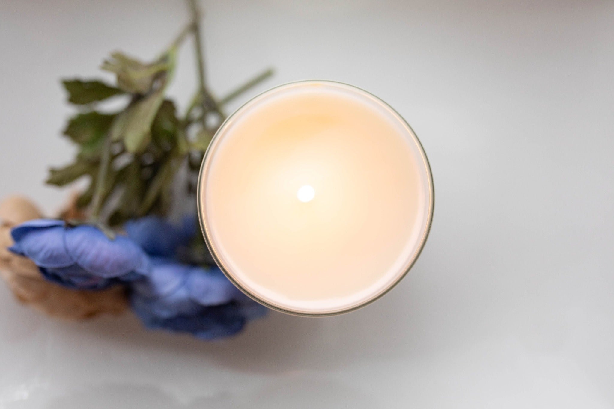 An overhead view of a single wick glass candle, lit, with a small bunch of flowers to the left of it.