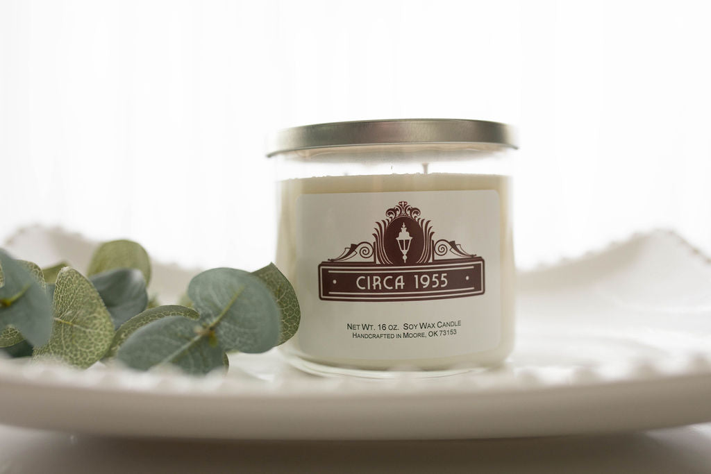 A three-wick clear glass candle with natural ivory wax sitting on a white hobnail glass tray.  On the left is a sprig of eucalyptus leaves.