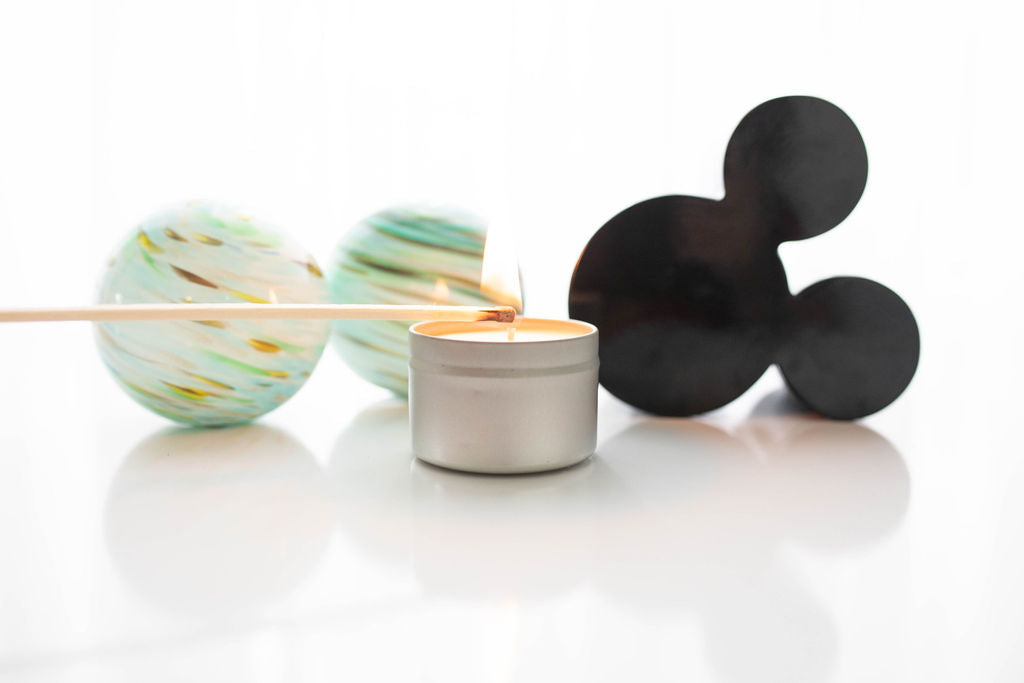 A silver candle tin with two decorative orbs to the left and a mickey head to the right.  The candle is being lit with a match.