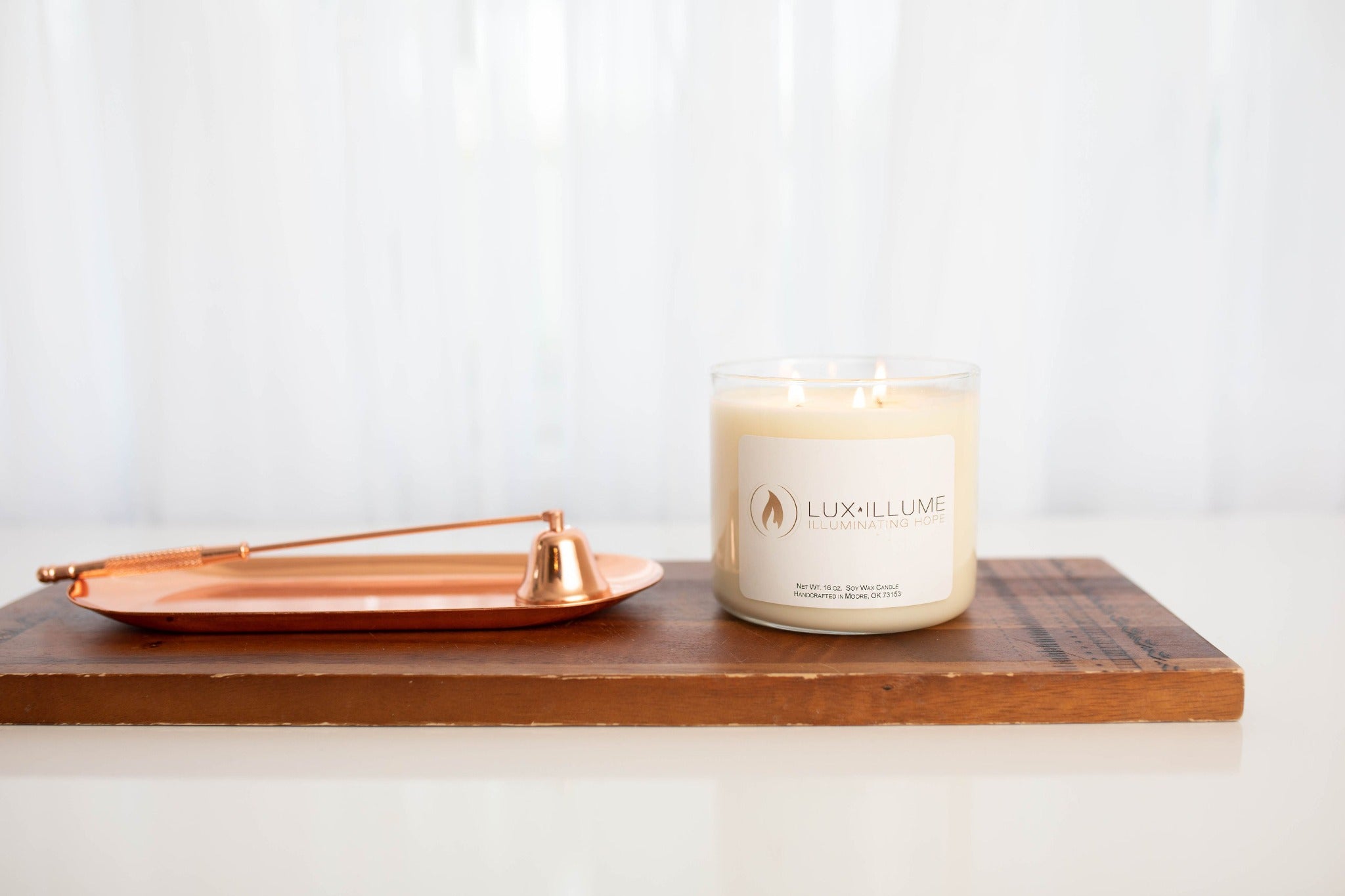 A three-wick clear glass candle with natural ivory wax.  It’s sitting on top of a wood block. Next to it on the left is a copper snuffer and tray.