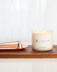 A three-wick clear glass candle with natural ivory wax.  It’s sitting on top of a wood block. Next to it on the left is a copper snuffer and tray.