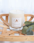 A single wick clear glass candle with natural ivory wax. The candle is in a wood slat bowl or tray with eucalyptus sprigs to the right of it.