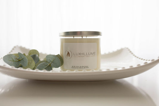  A three-wick clear glass candle with natural ivory wax sitting on a white hobnail glass tray.  On the left is a sprig of eucalyptus leaves.